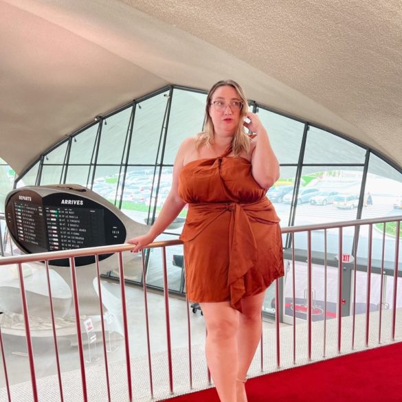 Sarah stands in copper one shoulder Gia IRL mini dress in the lobby of the TWA Hotel