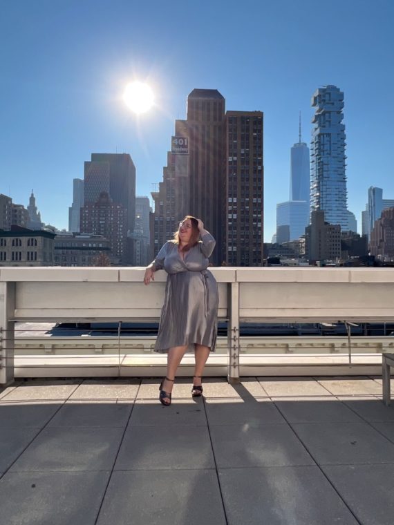 Image of Sarah standing on a rooftop in a silver dress with the sun and downtown NYC skyline in the background