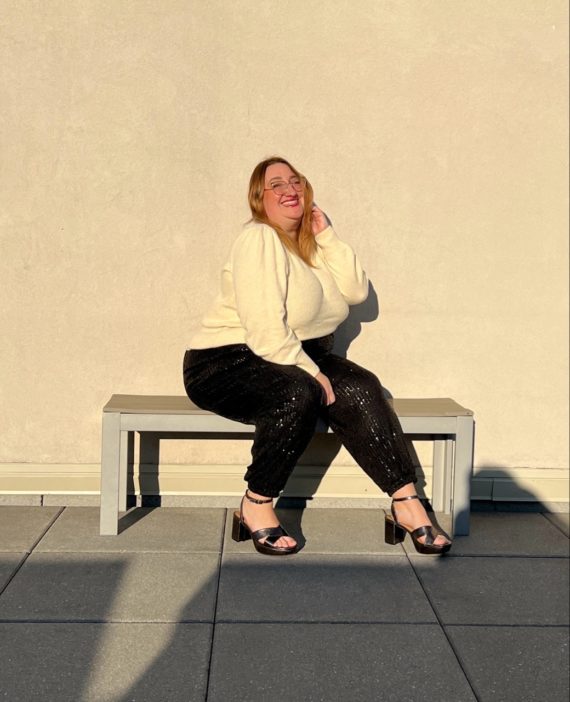 Sarah sits on a bench in the late afternoon sunlight in a cream sweater and black sequin joggers