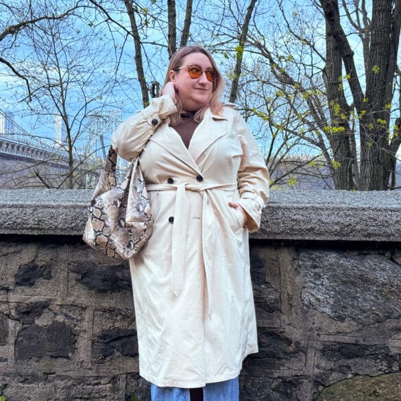 Sarah Chiwaya in a classic taupe trench coat standing in a park a dusk with a bridge in the background