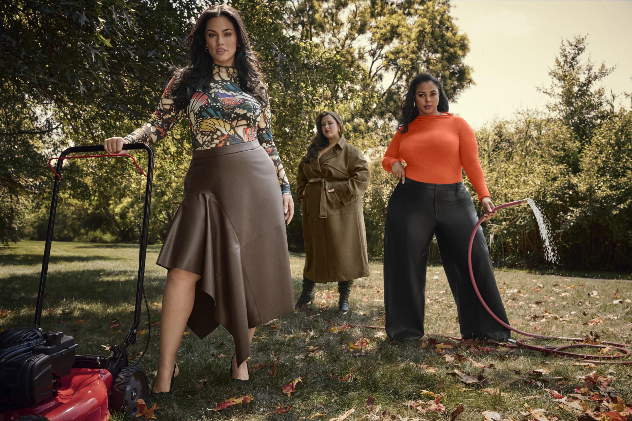 Ashley Graham Collection campaign image featuring three sharply dressed women in an outdoor scene with a lawnmower and a hose